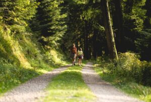 Two hikers walking a dirt trail through a dense, sunlit forest with tall trees on either side.